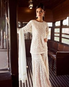 a woman standing in front of a window wearing a white dress with long sleeves and lace detailing