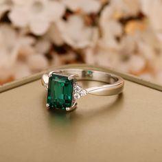 an emerald and diamond ring sitting on top of a gold box with flowers in the background