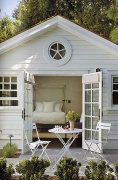 a small white house with a bed and table in the front yard, surrounded by greenery