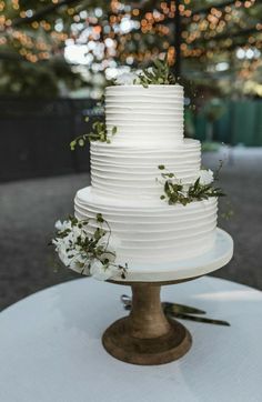 a white wedding cake sitting on top of a wooden stand with greenery and flowers