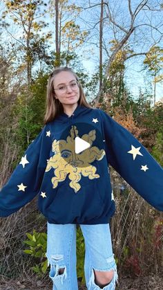 a woman wearing a blue hoodie with gold stars on it, standing in front of some bushes