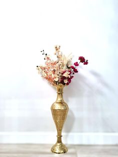 a gold vase filled with lots of flowers on top of a wooden floor next to a white wall