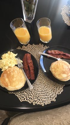 breakfast plates with eggs, sausages and pancakes on a black tablecloth next to two glasses of orange juice