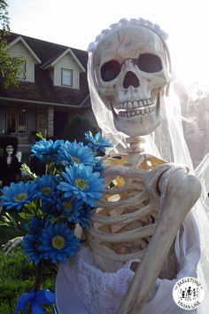 a skeleton dressed up as a bride in front of a house with blue flowers on the lawn