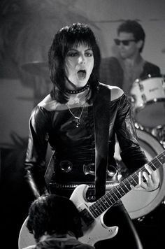 a woman with black hair playing an electric guitar in front of a man on stage