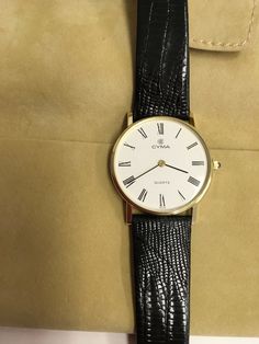 "Vintage round faced wrist watch made by Cyma.  Case is 14 karat yellow gold. It measures 30 mm in diameter and 5 mm thick.  The face is white with Roman numerals.  The stem has a small cabochon blue sapphire. The watch has a black leather strap measuring 9\".  Recently serviced.  The watch is preowned but in excellent condition.  Includes the original boxes and instructions." Gold Vintage Watch, Watch With Leather Strap, Roman Numerals, Wrist Watches, Vintage Watches, Blue Sapphire, The Face, Leather Straps, Wrist Watch