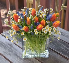 a clear vase filled with orange and white flowers on top of a wooden table next to a fence