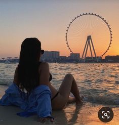 a woman is sitting on the beach looking at the ferris wheel in the distance with her back to the camera