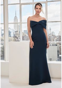 a woman standing in front of a window wearing a long dress with an off the shoulder neckline