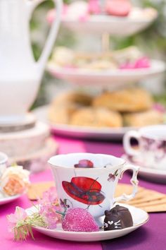 a table topped with plates and cups filled with pastries on top of purple cloth