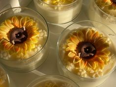 six sunflowers in small glass dishes on a white counter top, with the center surrounded by smaller yellow and orange flowers