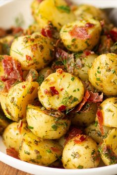 a white bowl filled with cooked potatoes covered in bacon and herbs on top of a wooden table