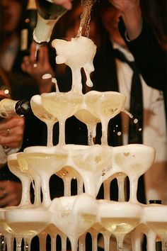 a group of people standing around a tall white fountain filled with wine glasses and champagne being poured into it