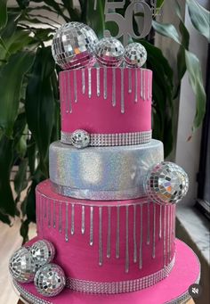 a pink and silver cake with disco balls on it's tiers is displayed in front of a plant