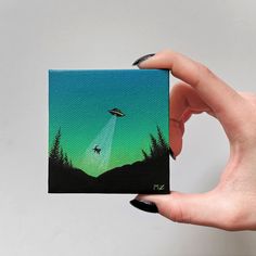 a hand holding up a small card with an alien flying in the sky above trees