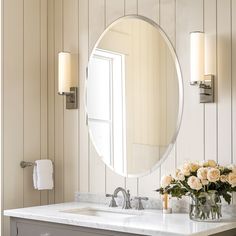 a bathroom vanity with a large round mirror above it and flowers in vase on the counter