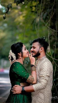 a man and woman standing next to each other in front of some green trees with their hands together