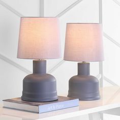 two gray lamps sitting on top of a white table next to a book and lamp shade