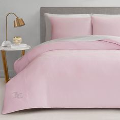 a bed with pink sheets and pillows in a room next to a lamp on a table