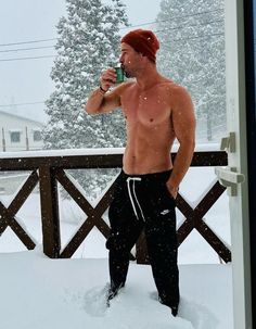 a shirtless man standing in the snow drinking from a cup and looking at his cell phone