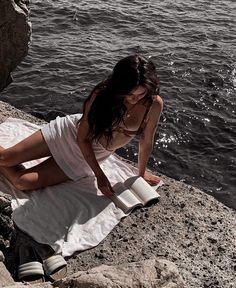 a woman sitting on the edge of a cliff reading a book by the water's edge