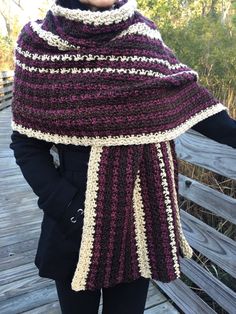 a woman wearing a purple and white crocheted shawl standing on a wooden bridge