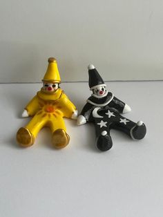 two toy clowns sitting next to each other on a white surface with one wearing a yellow hat