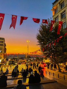 many people are sitting on the ground in front of some buildings and flags hanging above them