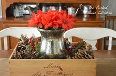 a silver vase filled with red flowers sitting on top of a wooden box covered in pine cones