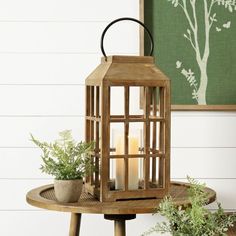 a wooden lantern sitting on top of a table next to potted plants and a framed tree