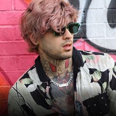 a man with pink hair and sunglasses standing in front of a graffiti covered wall, looking off to the side