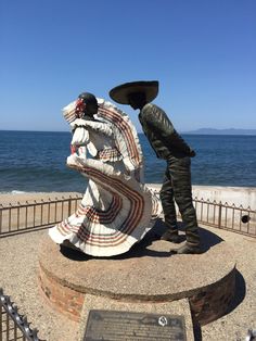 two people standing next to a statue near the ocean