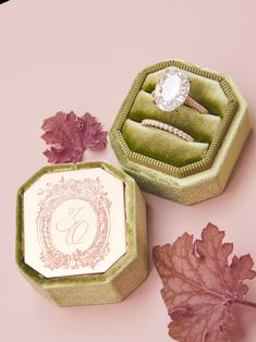 two wedding rings sitting in their boxes next to some leafy leaves on a pink surface