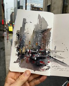 a person holding up an open book with watercolors on it in front of a city street