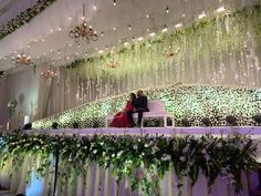 two people sitting on a bench in front of a stage with flowers and greenery