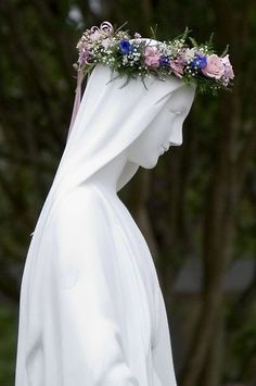 a white statue wearing a veil and flowers on it's head with trees in the background