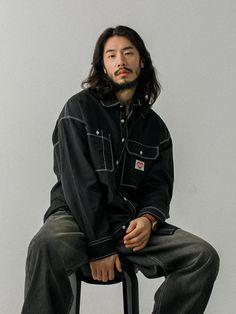 a man with long hair sitting on a stool and looking off to the side while wearing a black jacket