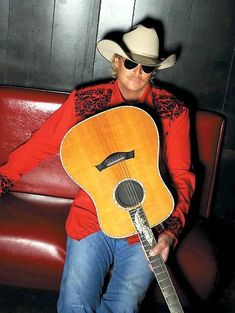 a man sitting on a red couch holding a guitar in his right hand and wearing a cowboy hat