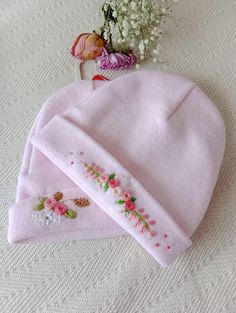 two pink knitted hats with embroidered flowers on the front and side, sitting on a white tablecloth