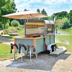 an outdoor food cart with stools and umbrella on the side of it, next to a body of water