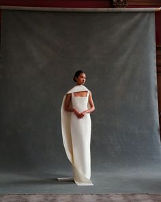 a woman standing in front of a gray backdrop wearing a white dress with cut out shoulders