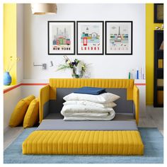 a yellow couch with pillows and blankets on it in front of two pictures hanging on the wall