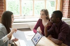 three people sitting at a table with laptops and papers in front of them, talking