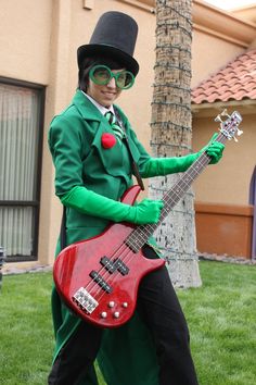 a man dressed in green and black holding a red guitar while standing next to a tree