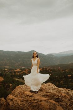 a woman in a white dress standing on top of a rock with mountains in the background