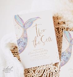 two mermaid themed wedding stationery cards on a woven basket with white and blue flowers