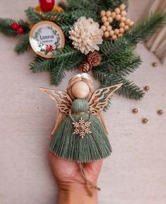 a hand holding a small green angel ornament next to pine cones and berries