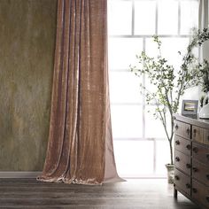 a room with a dresser, mirror and window covered in sheer drapes next to a potted plant