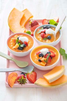 four bowls of fruit and cheese soup on a pink tray with spoons, slices of peaches, and orange wedges