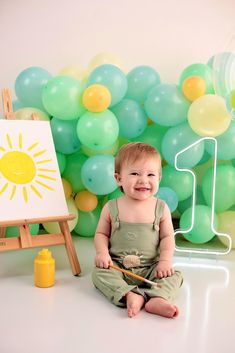 a baby sitting on the floor in front of balloons and an easel smiling at the camera